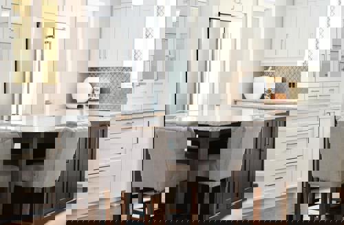 logo Bilden visar ett stilrent och ljust kök med vitmålade skåp och lådor, en köksö med mörkare trädetaljer och en marmorinspirerad bänkskiva. Framför köksön står två stoppade barstolar i grått tyg.