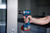 En person håller i en blå och svart borrmaskin med röd detalj, märkt "Bosch 18V Li-Ion" i ett rum.
