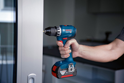 En person håller i en blå och svart borrmaskin med röd detalj, märkt "Bosch 18V Li-Ion" i ett rum.