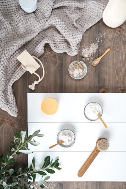 Grå filt, tvål, burkar och skedar med badsalt och krämer på träyta, med en växt i hörnet.