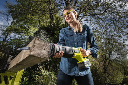 Person i skyddsglasögon använder en grön batteridriven tigersåg från Ryobi för att kapa trä utomhus.