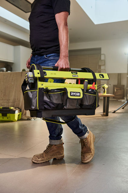 Person bär svart verktygsväska med gröna detaljer, märkta "RYOBI", i ett inomhusrum.