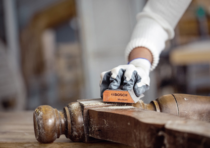 En person med vita handskar slipar en brun träyta med ett orange Bosch slippapper märkt "Medium".
