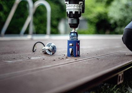 Borrmaskin med blå borrhålsåg som borrar i träplanka utomhus; lös träbit bredvid.