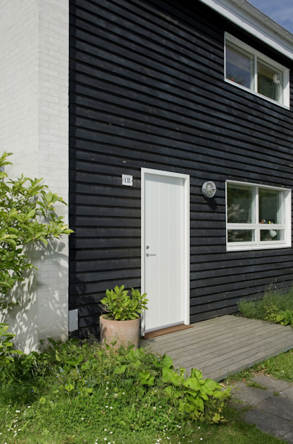 Svart träfasad med vit dörr och fönster; grönska och trätrall på marken.