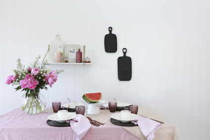 Dukat bord med rosa duk, rosa servetter, blommor, vattenmelon, glas och två svarta skärbrädor på vit vägg.