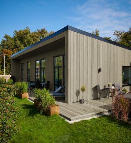 Modernt envåningshus med träterrass, stora fönster och grönskande omgivning.