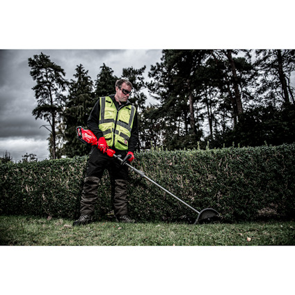 Man in a high-visibility vest using a red trimmer on a hedge, trees and cloudy sky in the background.