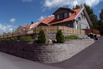 Hus med rött tak och grå fasad, omgiven av trästaket och stenmur, på upphöjd tomt med asfalterad infart.