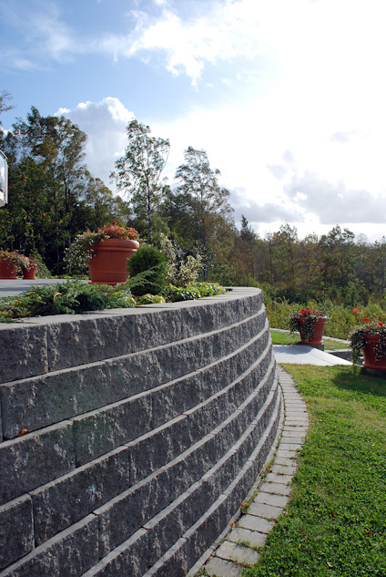 Murar med grå staplade stenar, krukväxter ovanpå, omgiven av gräs och träd.