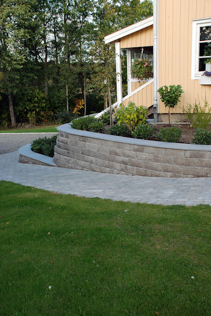 Hus med gul fasad och vita fönster, stenlagd uppfart och en stödmur med växter framför. Gräsmatta i förgrunden.