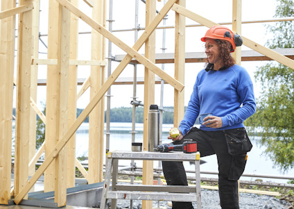 Man med bygghjälm och hörselskydd håller en frukt och mugg nära byggställning vid sjö.