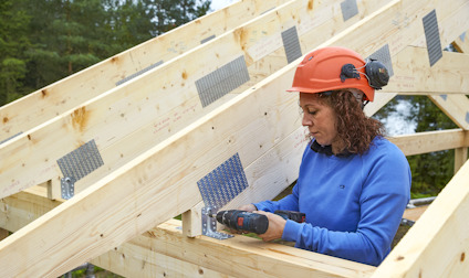Person i blå tröja och orange hjälm som arbetar med träkonstruktion, håller en skruvdragare.