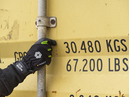 Hand med svarta handskar håller i metallstång framför en gul container med texten "30,480 KGS 67,200 LBS".