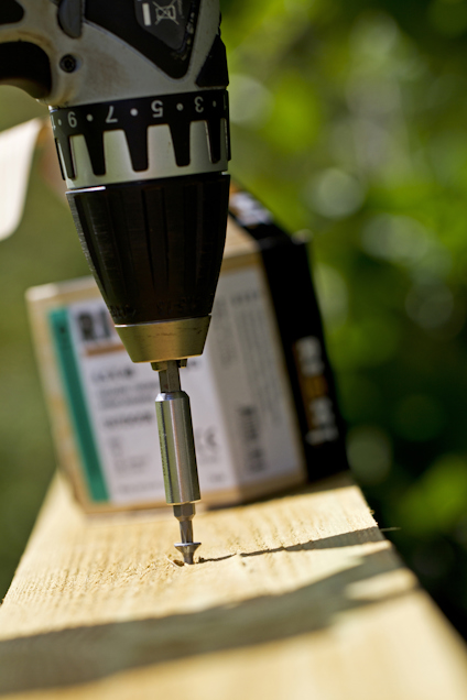 Borrskruvdragare skruvar en skruv i ett trästycke utomhus. En kartong syns i bakgrunden.