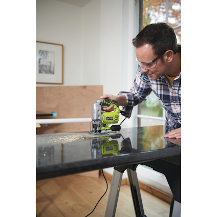 Person använder grön sticksåg på svart bordsskiva, med skyddsglasögon och skjorta i ljus miljö.