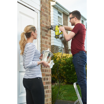 Man använder borrmaskin för att fästa något på tegelvägg; kvinna håller lampa.