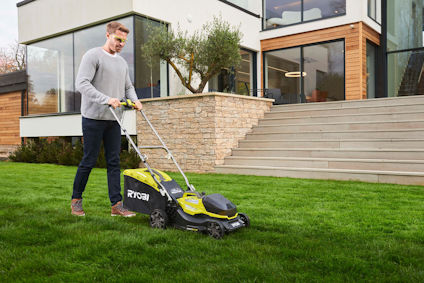 Person använder en RYOBI gräsklippare på en grön gräsmatta framför ett modernt hus.
