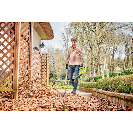 Person med rutiga kläder använder en lövblåsare för att flytta löv på en grusgång bredvid en trätrall.