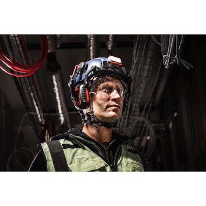 En person i skyddsutrustning, inklusive hjälm, skyddsglasögon och väst, står inomhus med industriledningar i bakgrunden.