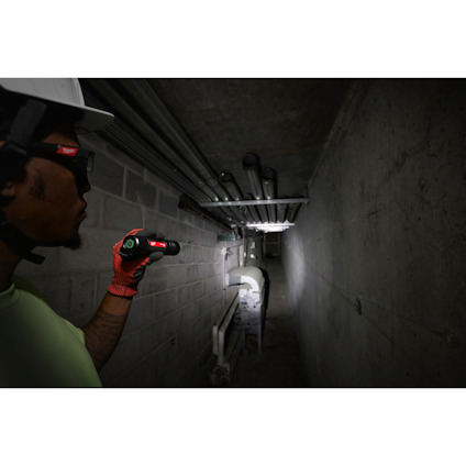 Person med hjälm och handskar lyser med ficklampa i betongtunnel med rörledningar.