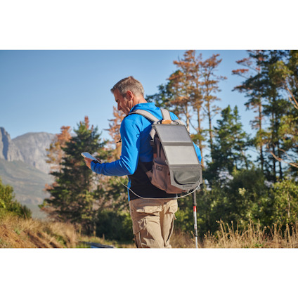 En person i vandringskläder med ryggsäck använder en smartphone på en skogsstig, omgiven av träd och berg i bakgrunden.