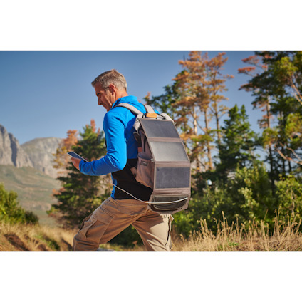 Person i blå jacka och beige ryggsäck går på en stig med träd och berg i bakgrunden.