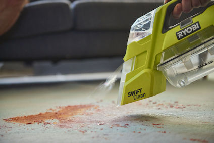 En handhållen dammsugare i grön färg från märket Ryobi städar ett spill på en matta.
