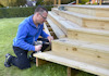 Man kneeling on grass using a drill on wooden outdoor steps, with screws and tools nearby.
