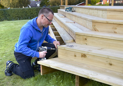 Man kneeling on grass using a drill on wooden outdoor steps, with screws and tools nearby.