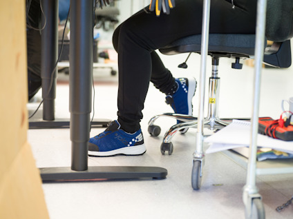 En person i svarta byxor och blåa sneakers sitter på en kontorsstol vid ett skrivbord.