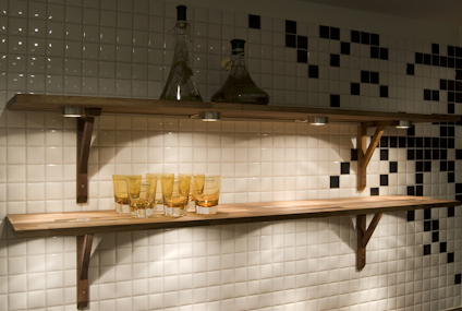 Två trähyllor monterade på en vit och svart kaklad vägg, med glasflaskor och glas på hyllorna.