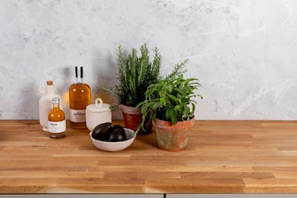 Kryddväxter i terrakottakrukor, avokado i en skål, och flaskor på en träbänkskiva mot en ljus stenvägg.