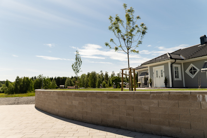 Tegelstensmur med träd och beskurna buskar framför ett hus med grått tak i soligt landskap.