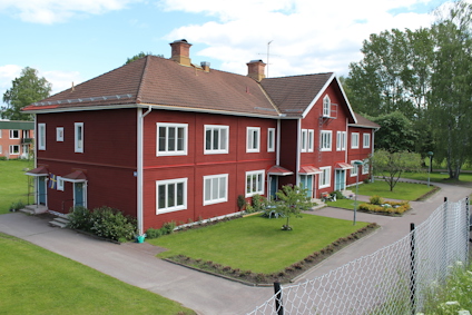 Rött tvåvåningshus med vita fönsterkarmar, omgiven av trädgård; träden i bakgrunden.