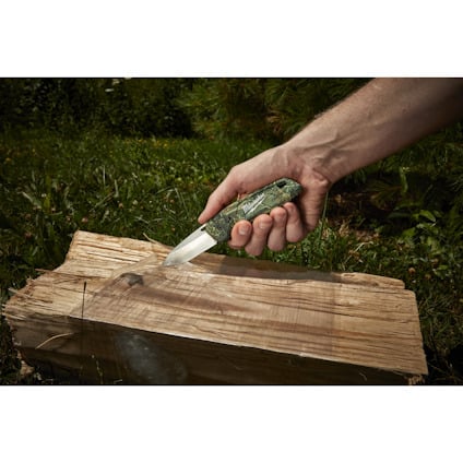 Hand skaver trä med en kniv med grön mönstrat handtag över en trästock i en skogsmiljö.
