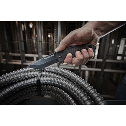 En hand håller en svart fällkniv ovanför en spole med elektriska rör i en industriell miljö.