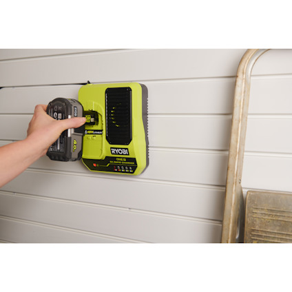 Hand placerar ett batteri i en grön snabbladdare av märket RYOBI monterad på en vägg med vita paneler.