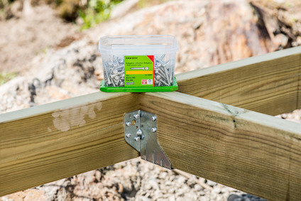 Plastlåda med skruvar och grön etikett med texten 'Ankarskruv', placerad på träreglar med metallbeslag utomhus.