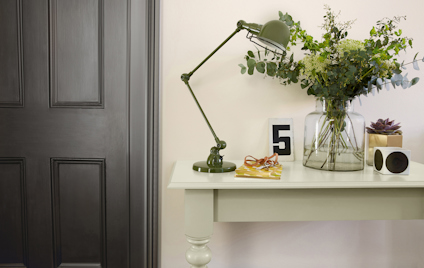 Grönt sidobord med bordslampa, glasvas med blommor, böcker och en mörk trädörr i bakgrunden.