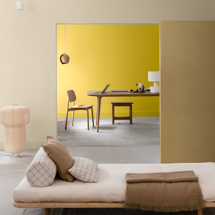 Modernt vardagsrum med gul accentvägg, träbord och stol i bakgrunden, beige soffa med kuddar i förgrunden.