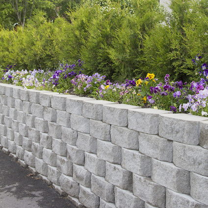 Grå mur av staplade stenar med blommor ovanpå, gröna buskar i bakgrunden.