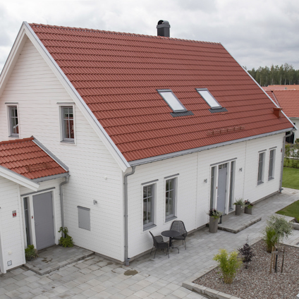 Vit träfasad med rött sadeltak, två takfönster. Marksten runt huset, trädgårdsmöbler vid entrén.