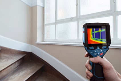 Händer som håller en värmekamera, pekar mot ett fönster i ett rum med trappa. Skärmen visar en termisk bild.