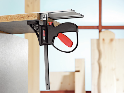Skruvtving med rött handtag fäster trästycke vid kant på bänkskiva. Bakgrund med träramar och trästycken.