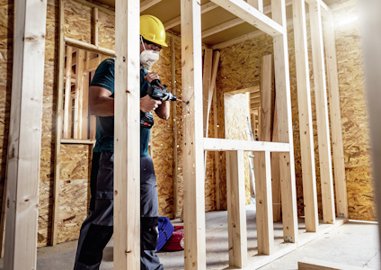 Byggnadsarbetare med hjälm och skyddsmask använder borrmaskin på träkonstruktion.