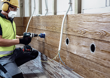 Person i skyddsutrustning borrar hål i en träpanel inomhus med en elektrisk borrmaskin.