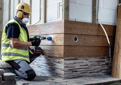 Person med skyddsutrustning borrar i träpanel vid en vägg.