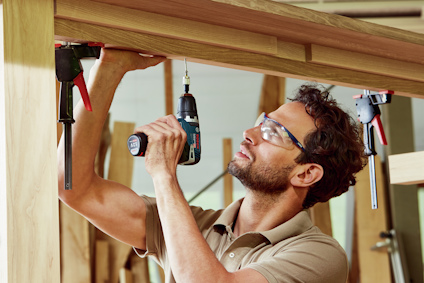 Man använder en bärbar elektrisk borr för att fästa trästycken med hjälp av klämmor, iförd skyddsglasögon.