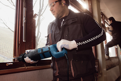 Person som använder en blå elektrisk såg för att skära i metall eller trä vid ett fönster. Ytterligare person syns i bakgrunden.